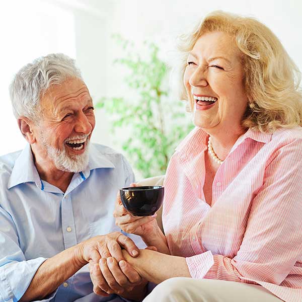 An elderly man and woman sit close together, laughing joyfully. The woman holds a coffee cup, and they both appear happy and relaxed in a bright, cozy room with greenery in the background.
